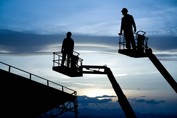 Two construction workers rise to the top of a metal framed warehouse.
